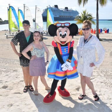 Girl and women with Minnie Mouse at tropical beach, sailboats, cruise ship in background, relaxing vacation scene.