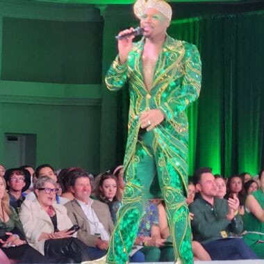 Drag performer in green and gold costume on stage at Disney fashion event.