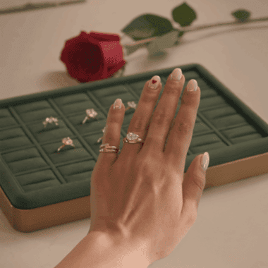 Hand with engagement ring and wedding bands on a jewelry tray with a red rose.